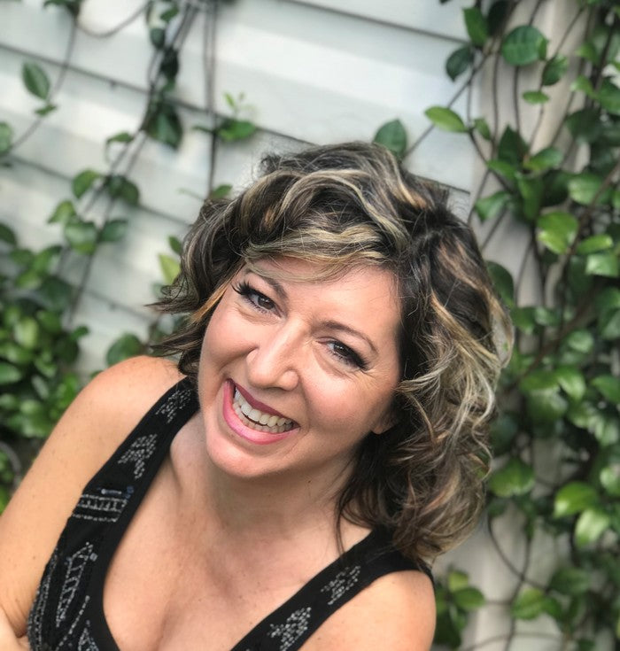 Heather Burch is a middle-aged white woman with curly, shoulder-length, dark hair with light highlights. She has a broad smile. She is photographed in front a white wall covered with greenery.