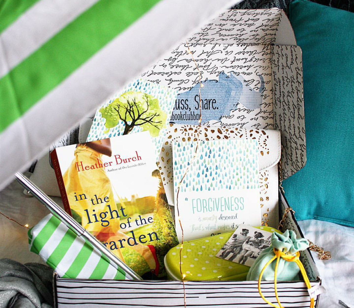 A paperback copy of In the Light of the Garden by Heather Burch sits inside an open Once Upon a Book Club box surrounded by the unwrapped gifts inspired by the book including a green and white striped umbrella, a strand of fairy lights, a yellow polka-dotted container, a photograph, and a bag of soothing mud mask (recipe in the back of the book).