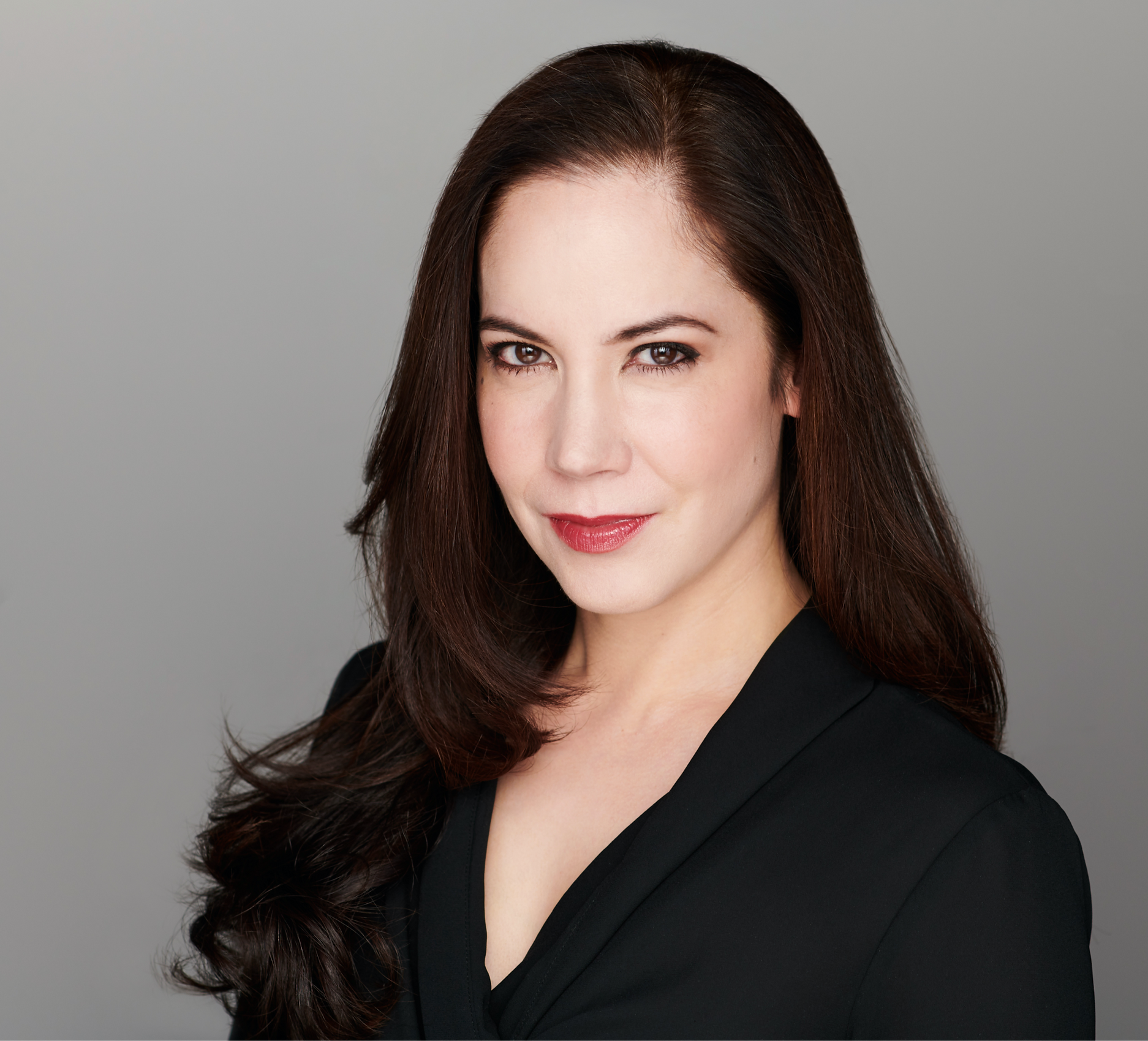 Author Karin Tanabe is a young white brunette woman standing in front of a gray background wearing a black dress