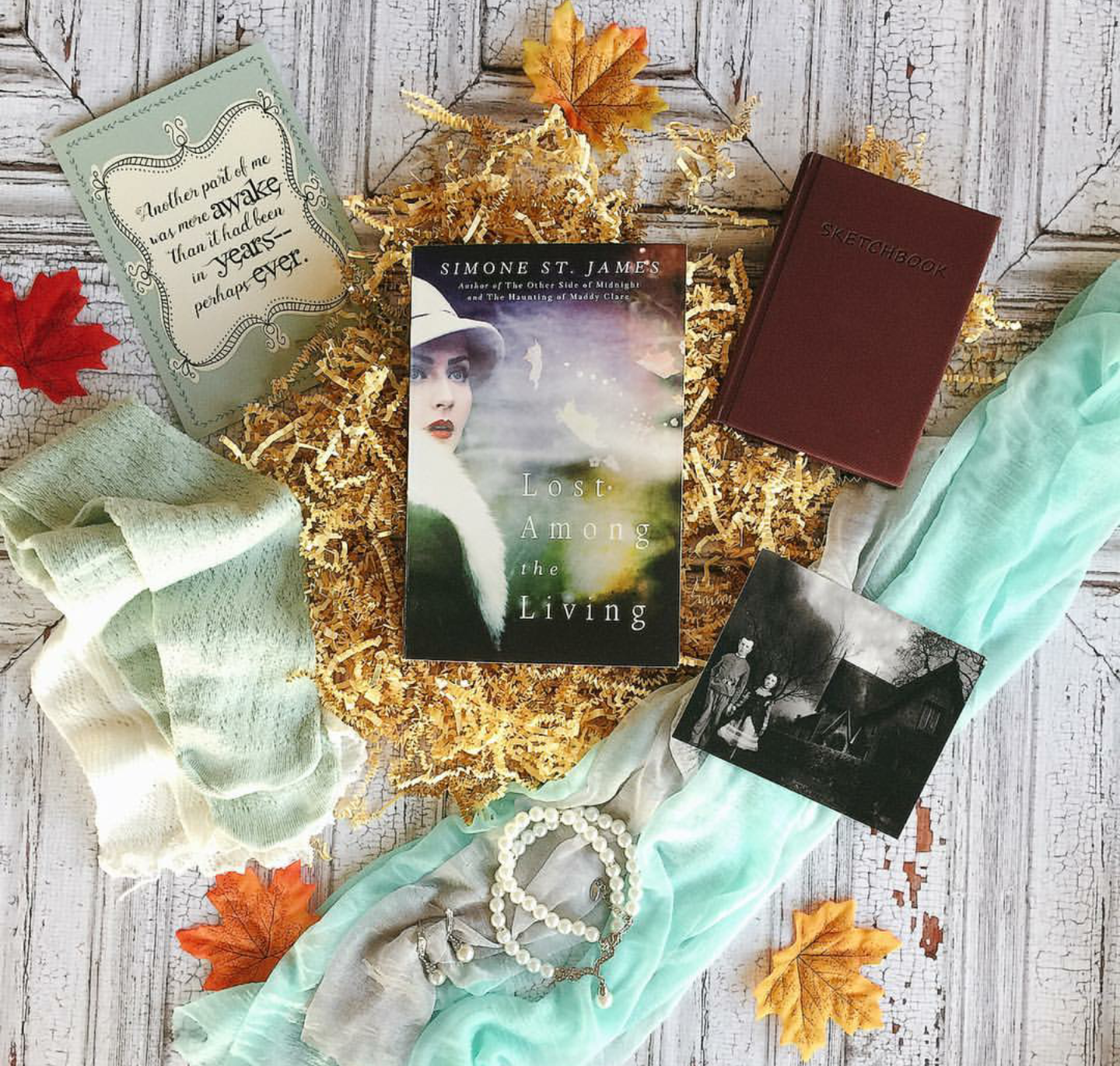 A paperback copy of Lost Among the Living by Simone St. James sits on wood floor surrounded by crinkle paper and faux autumn leaves. Also featured is the quote print, a pair of socks, a photograph, a sketchbook, a scarf, and a pearl necklace with earrings.