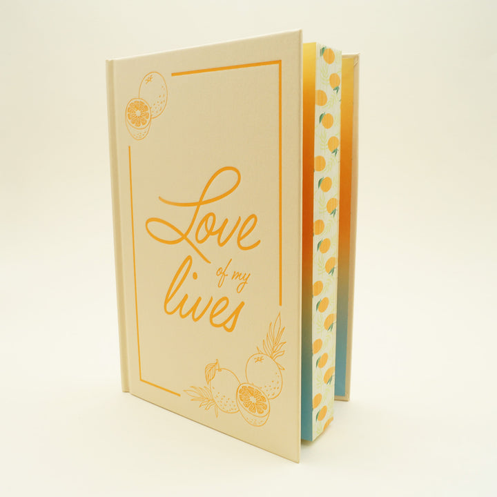 a hardcover book "Love of my Lives with images of oranges on the top left and bottom right stands slightly open, showing a pattern of oranges printed on the pages