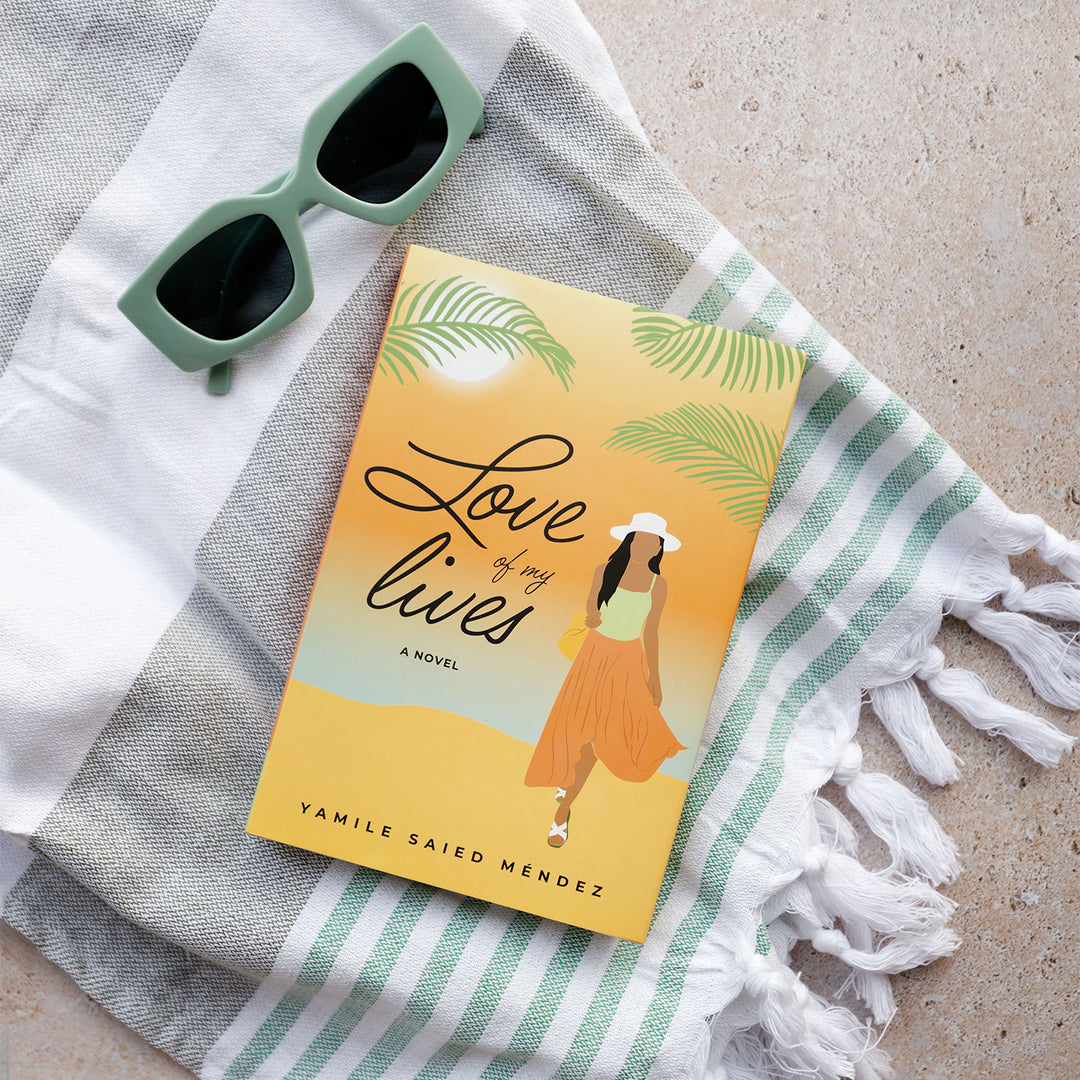 a hardcover copy of "Love of my Lives" by Yamile Saied Méndez sits on a beach towel next to green sunglasses. On the cover, there is a beach setting and a woman faces forward in an orange skirt and green top with a white sunhat