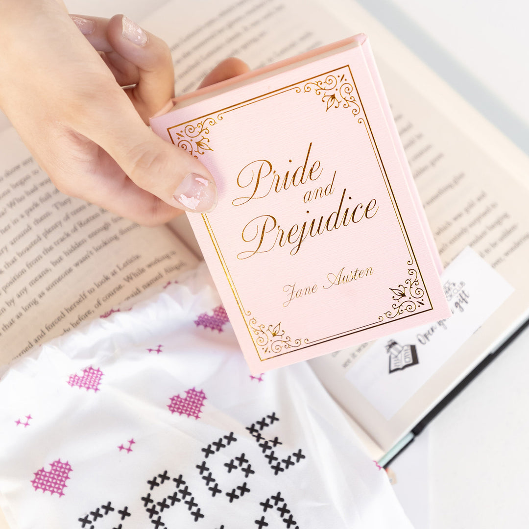 A white hand holds a mini pink jewelry box labeled Pride and Prejudice Jane Austen in gold lettering over an open book with an open your gift sticky note at the bottom of the page.