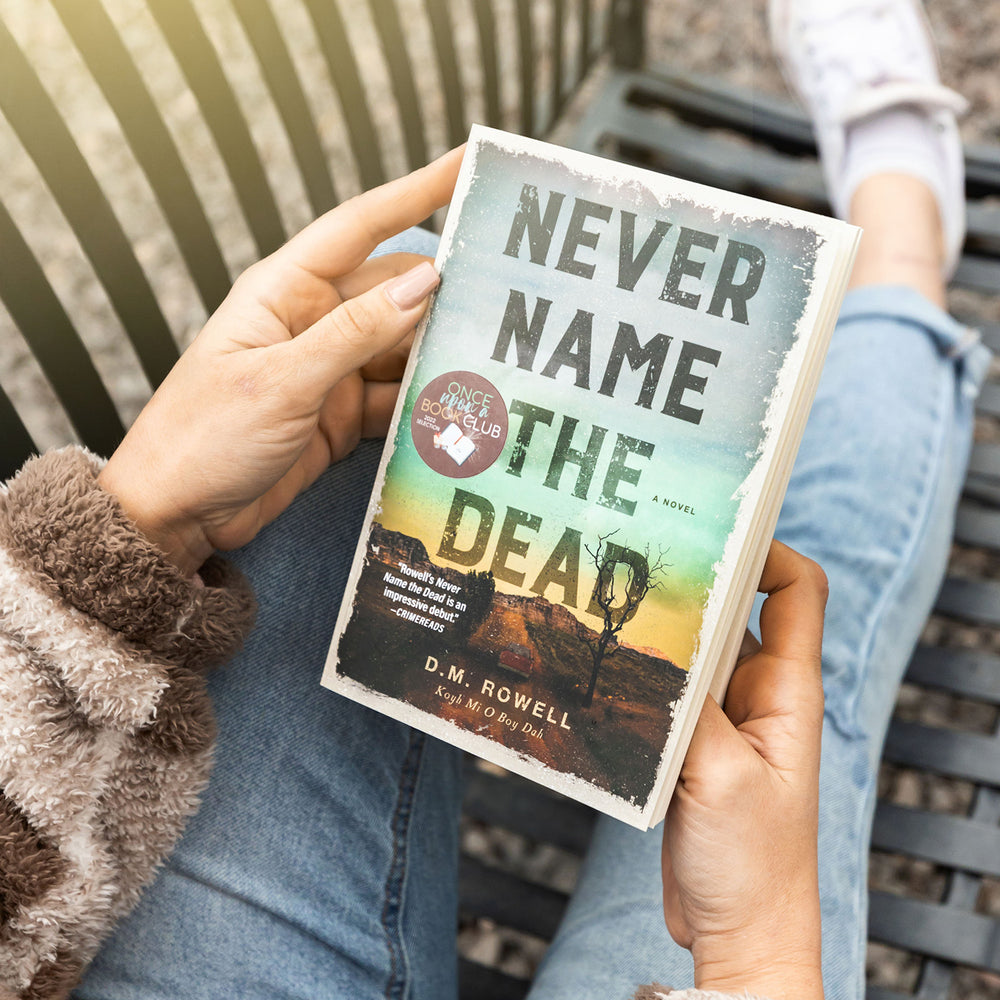 a white woman holds a paperback edition of Never Name the Dead by DM Rowell while sitting on a park bench