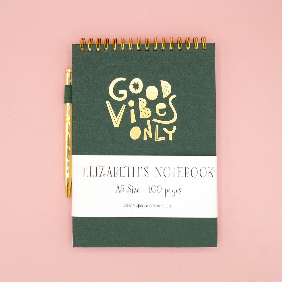 A dark green spiral notebook that says Good Vibes Only in gold lettering on the front. A gold pen is attached to the side.