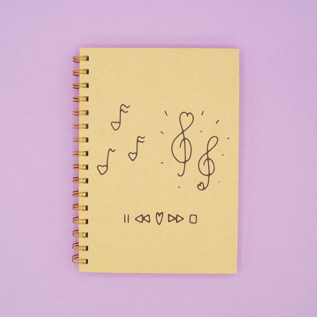 a light brown hard cover notebook with music notes drawn on the front cover sits on a purple background.