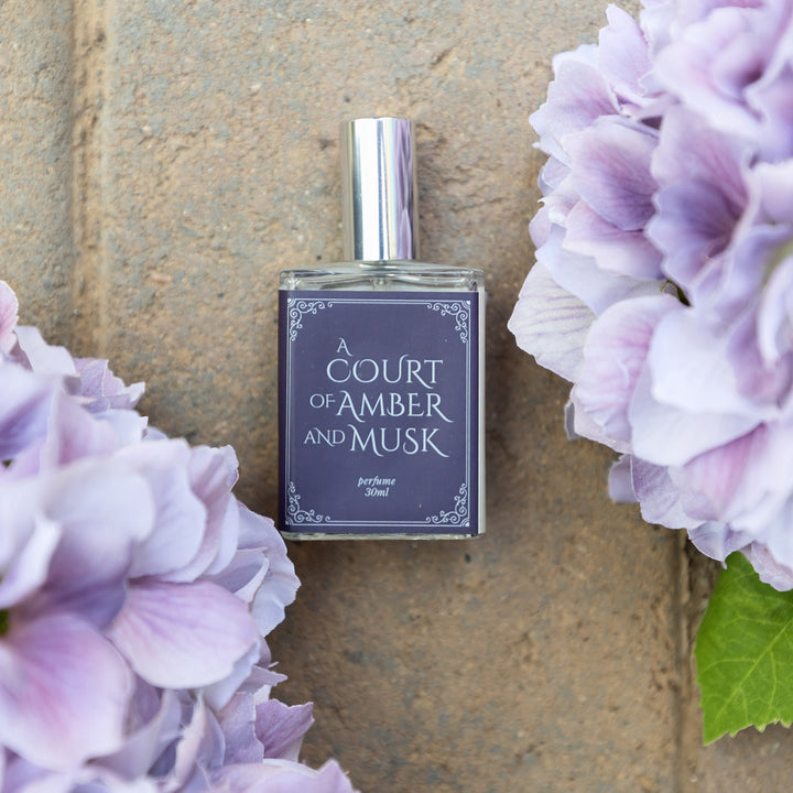 A close-up image of the bottle of perfume scented "A Court of Amber and Musk" showing the dark and light blues of the bottle's label. The bottle lays on a cement brick background surrounded by purple flowers.