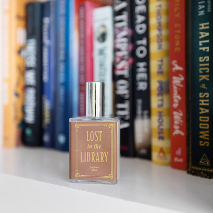 A close-up image of the bottle of perfume scented "Lost in the Library" showing the light brown and gold toned writing on the label. The bottle sits on a bookshelf in front of multiple titles.