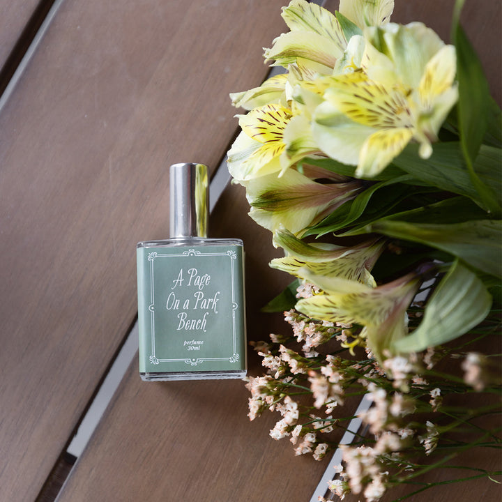 A close-up image of the bottle of perfume scented "A Page on a Park Bench" showing the light green and white details on the label. The bottle lays on wood slat background next to a small bunch of various flowers.