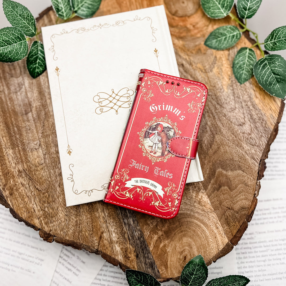 Grimm's Fairy Tales red clutch with gold accents on a wooden surface surrounded by green leaves.