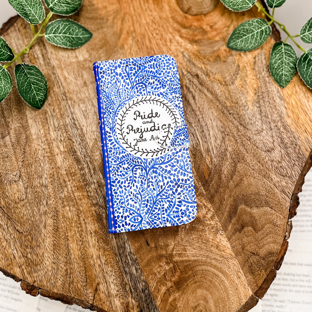 Pride and Prejudice-themed clutch with a blue floral design on a wooden surface with green leaves.