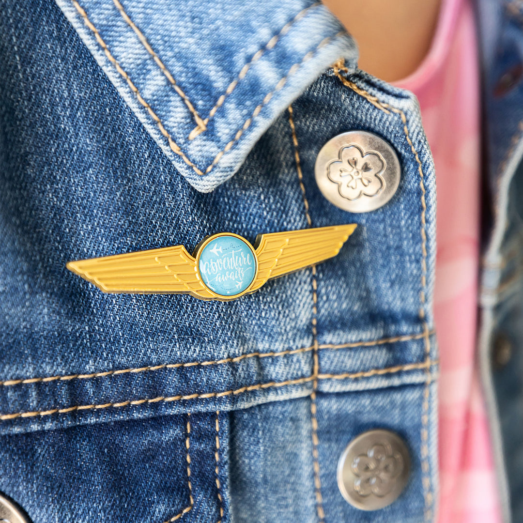 an enamel pin with gold wings and a blue circle at the center that says "Adventure Awaits" is pinned to the front of a person's jean jacket