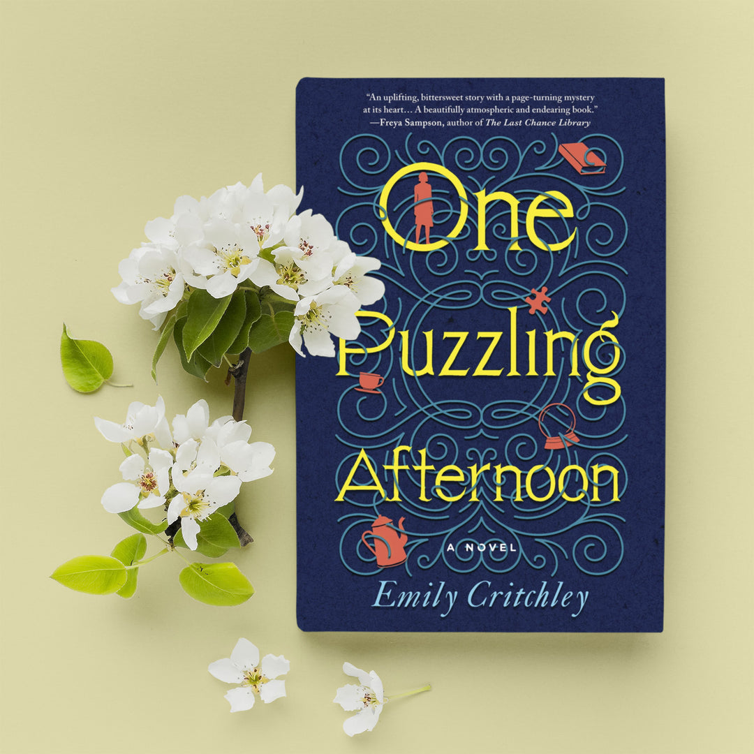 A paperback copy of One Puzzling Afternoon by Emily Critchley. White flowers are next to the book.