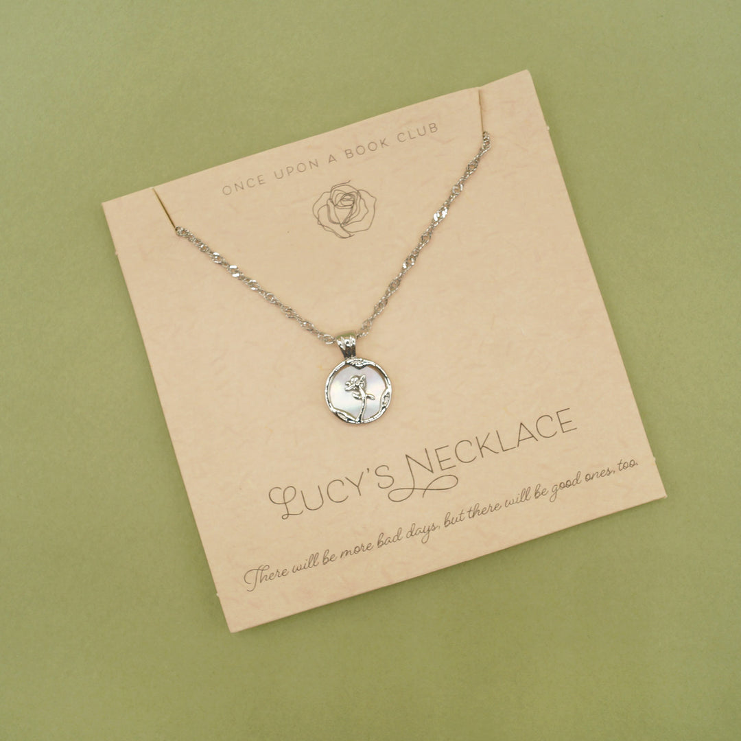 A small silver pendant with a rose sits on necklace card. The words "Lucy's necklace" is written along the bottom of the card.
