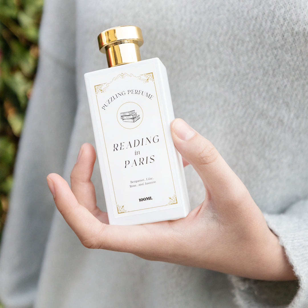 "Reading in Paris" perfume. A white squared off jar with a gold lid. Held by a white hand in front of a grey sweater.