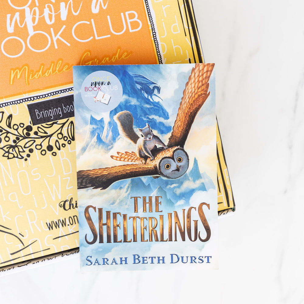 A close-up of the paperback copy of The Shelterlings by Sarah Beth Durst sitting on top of a yellow Once Upon a Book Club box.