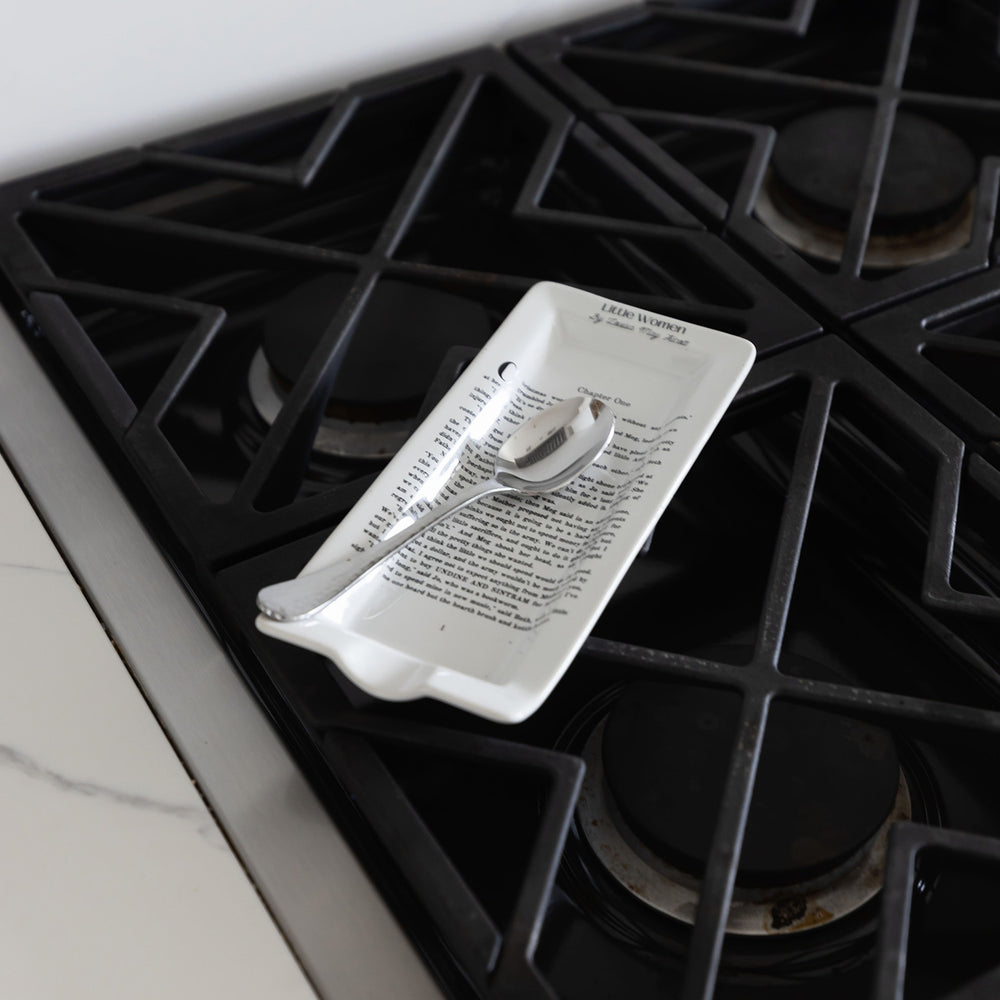 Literary-themed spoon rest with text design, holding a silver spoon, placed on a black stovetop.