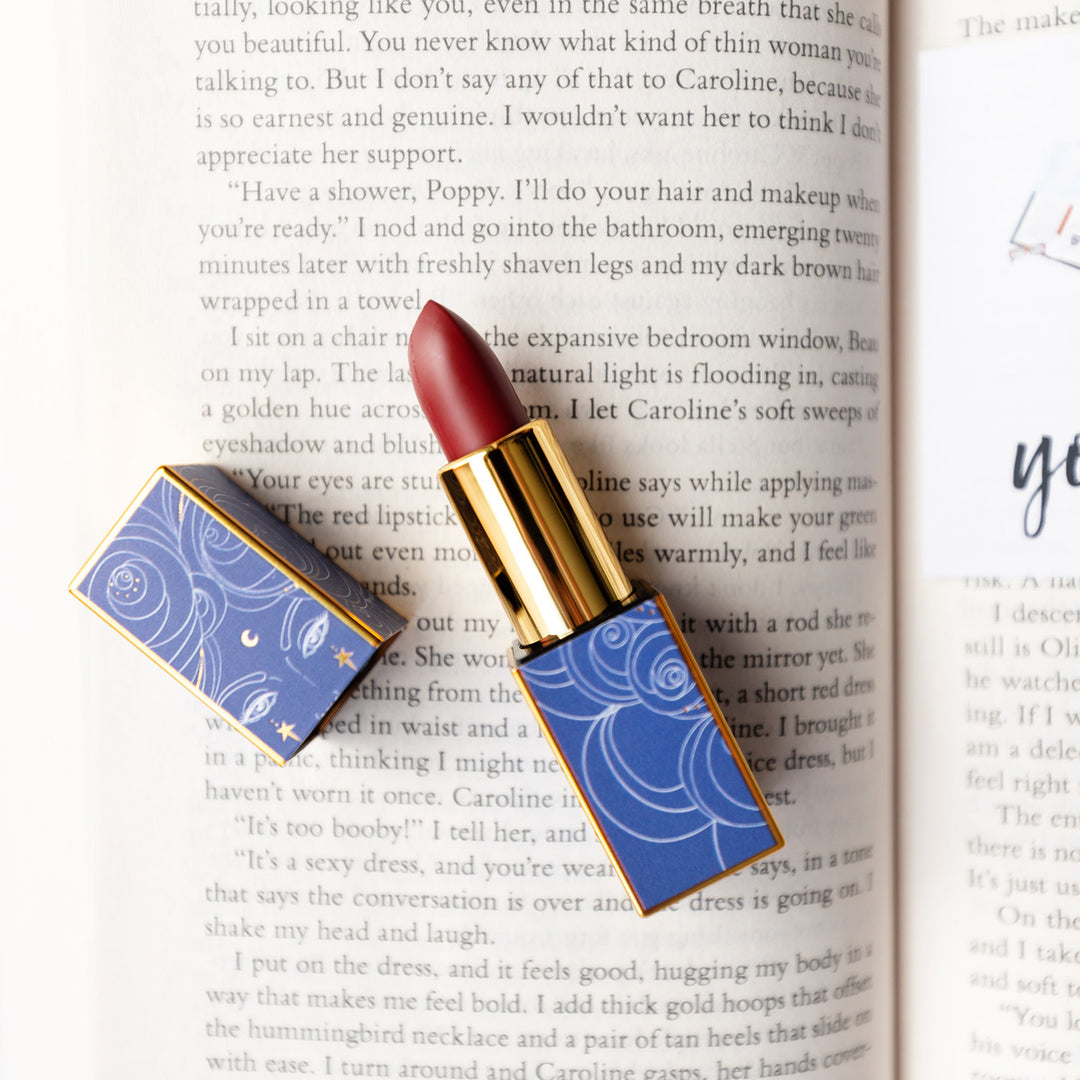 A stunning, deep red shade of lipstick in a beautiful blue and gold, squared, magnetic tube laying open on an open book.