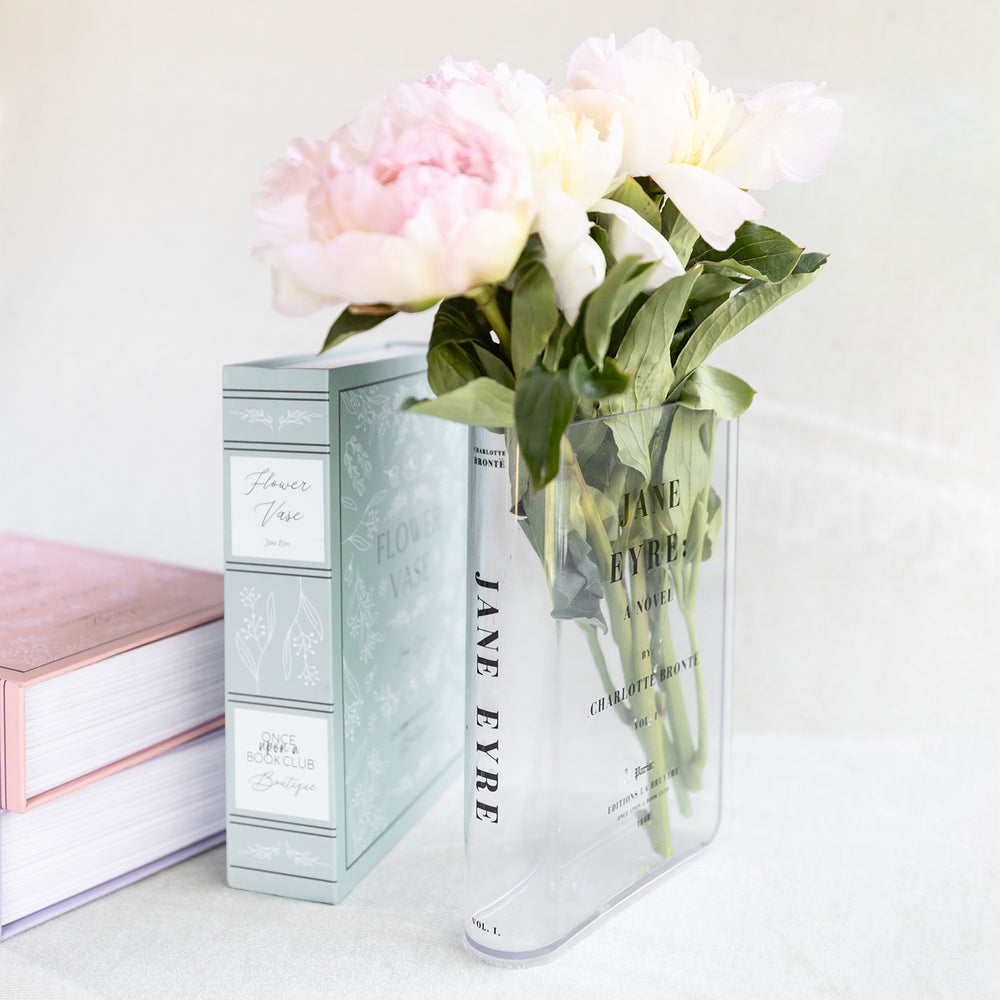 Clear vase with "Jane Eyre" design holding pink peonies, beside its decorative box and books.