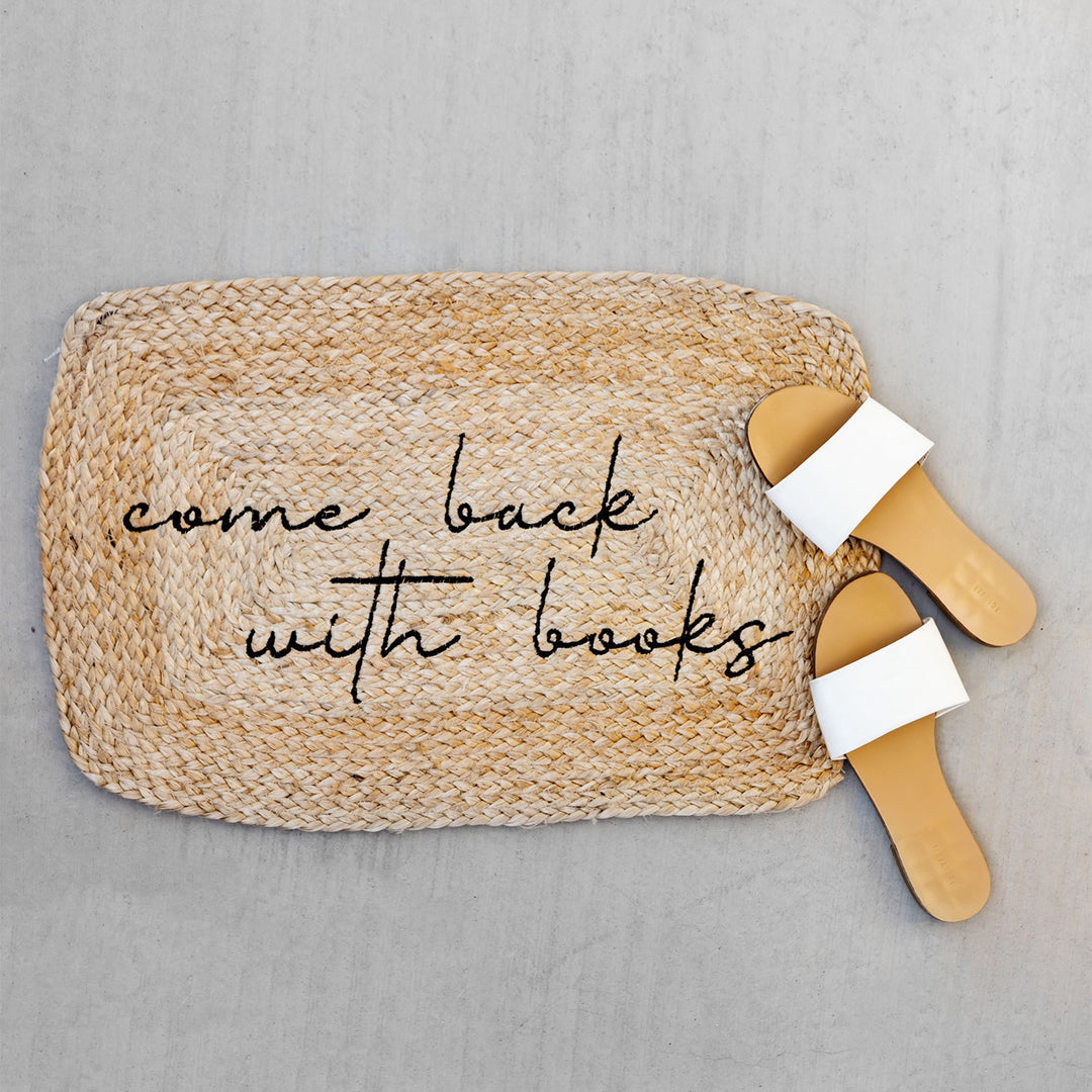 a jute welcome mat embroidered with black thread with the phrase Come Back with Books and a pair of beige and white sandals on the right edge of it