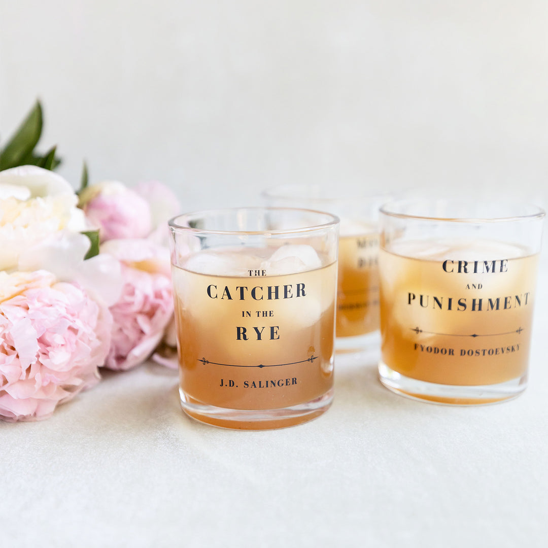 Three whiskey glasses featuring classic book titles, surrounded by soft pink and white flowers.
