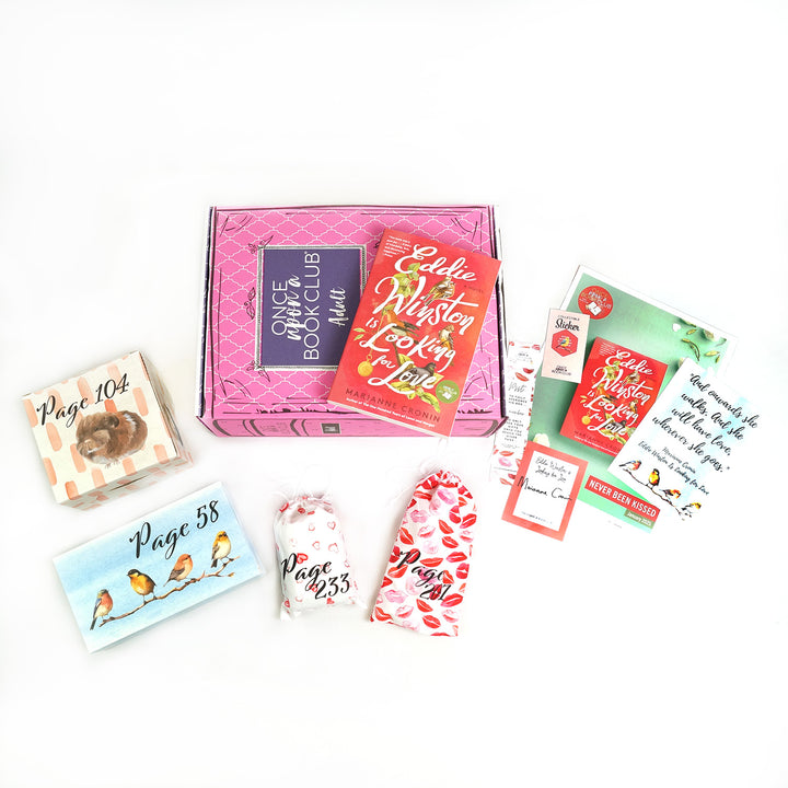 Paperback edition of Eddie Winston is Looking for Love lays on a pink OUABC box. Also included are four themed wrapped gifts labeled with page numbers and paper items.