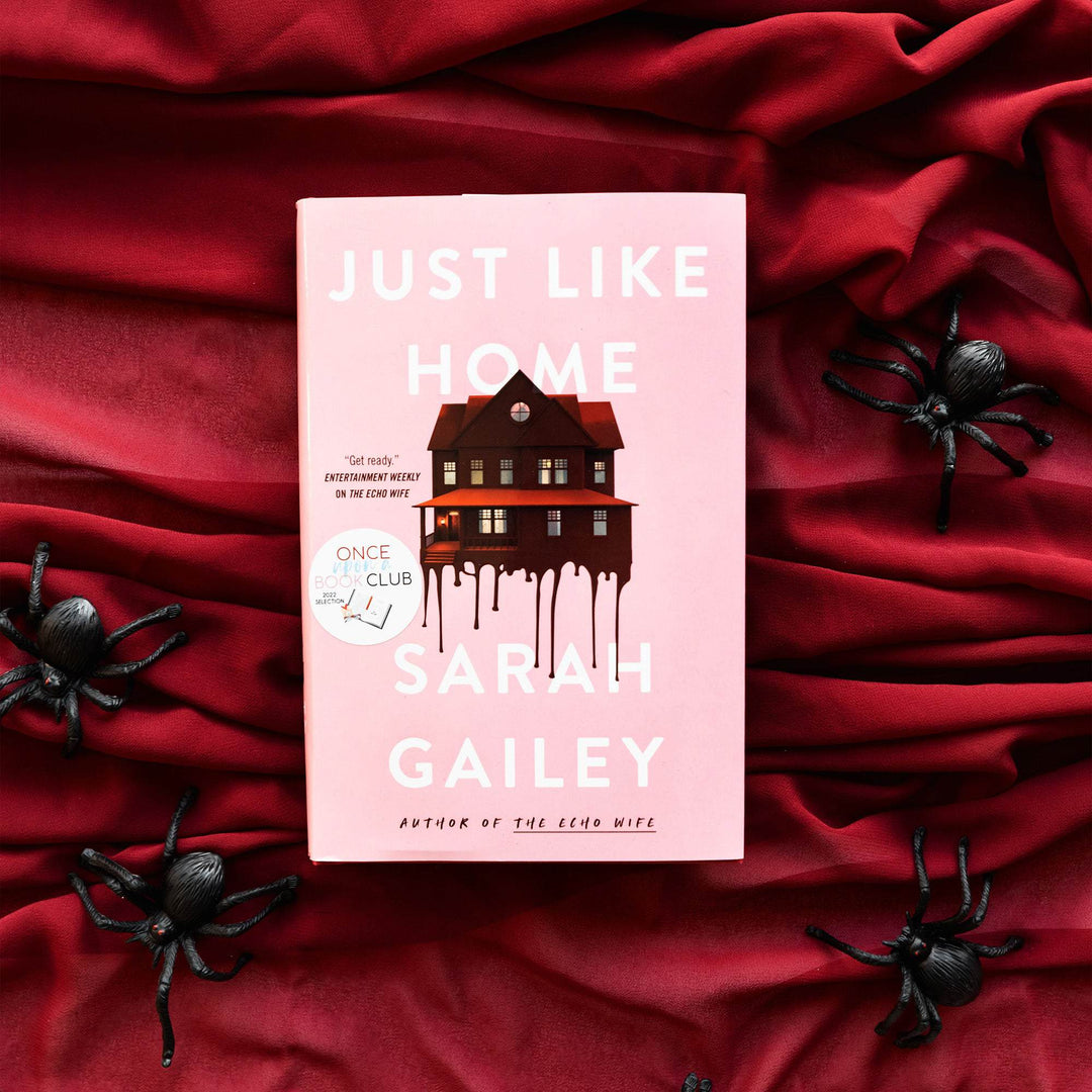 a hardcover edition of Just Like Home lays on a red blanket surrounded by fake black spiders