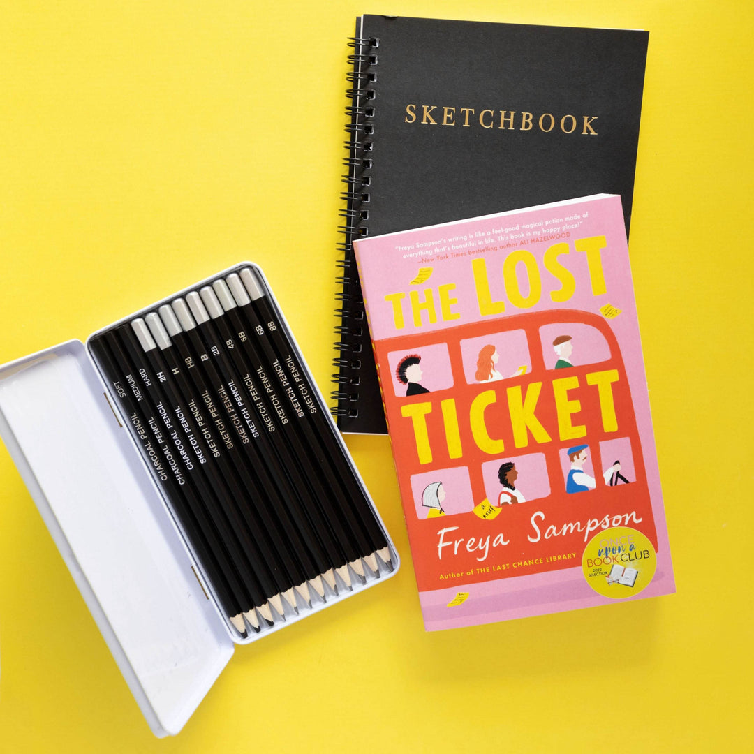 a paperback edition of The Lost Ticket by Freya Sampson sits on a black sketchbook next to a set of black pencils in a white case