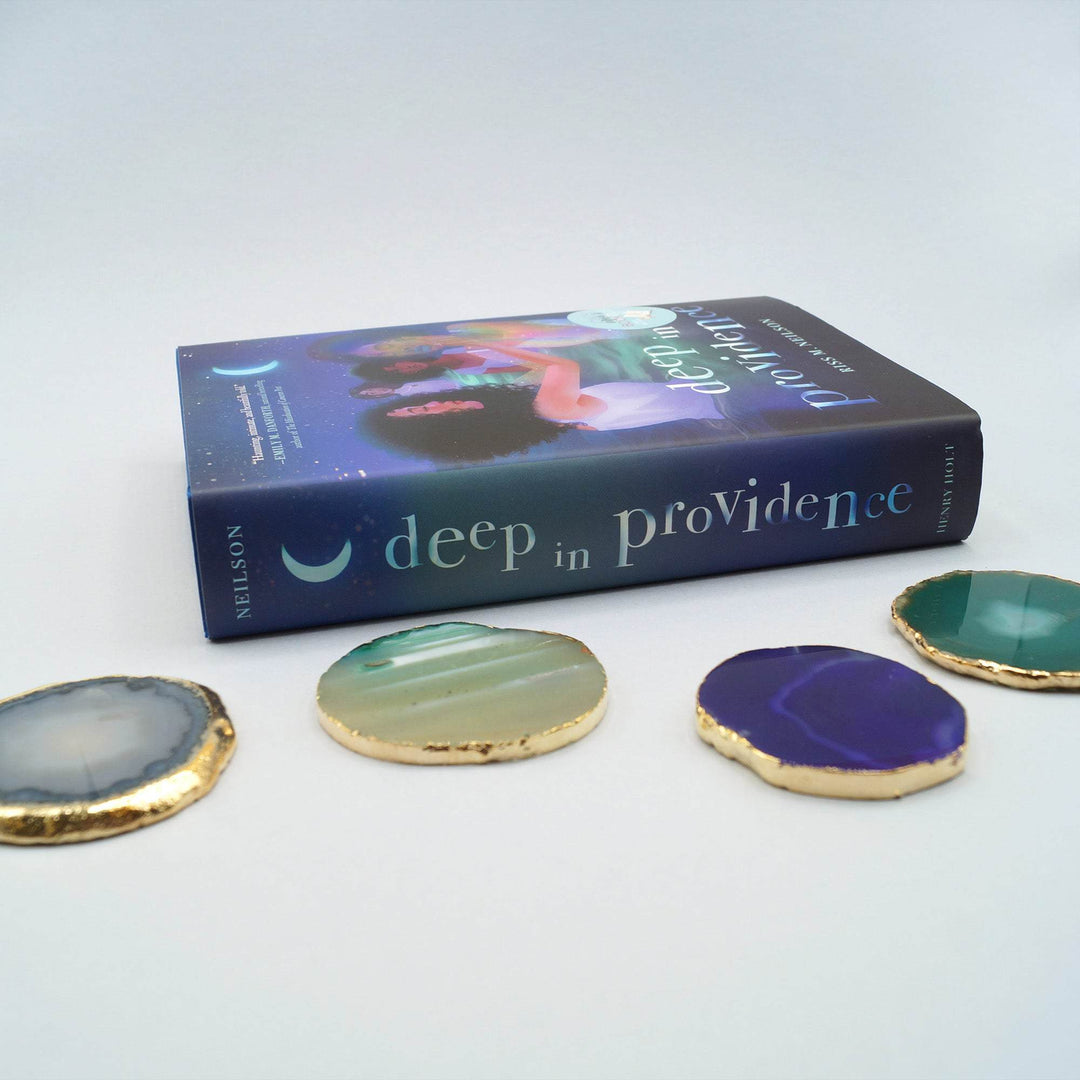 a hardcover edition of Deep in Providence lays on its side. In front of it are four agate geode coasters in gray, green, and purple with gold lining each one