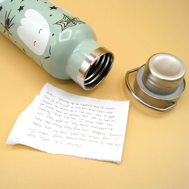 Open green water bottle with ghost designs, a removed lid, and a handwritten note on yellow surface.