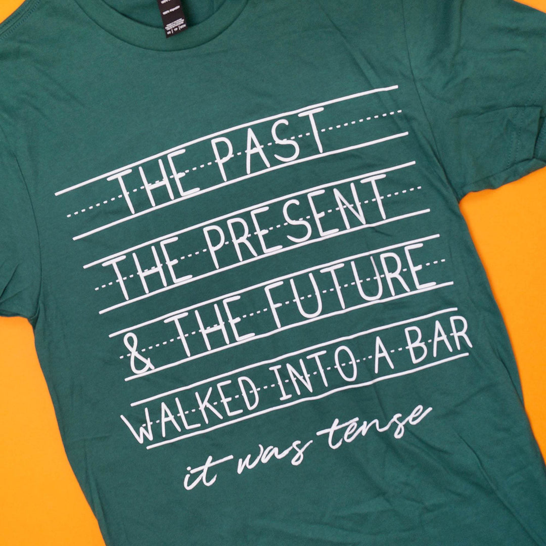 a green t-shirt with the quote "The Past, The Present, & The Future walked into a bar. It was tense" in white writing