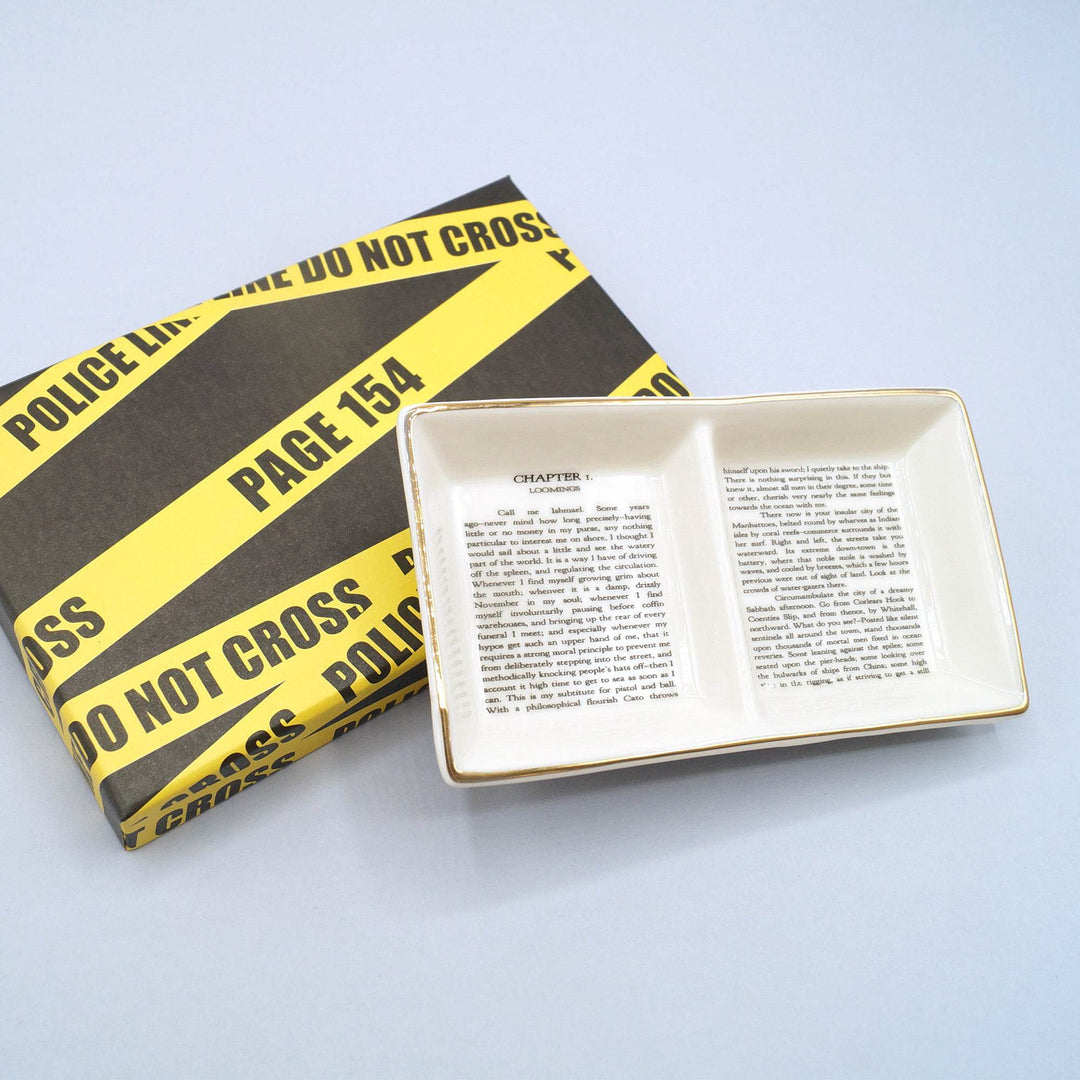Ceramic ring dish printed with book text, placed beside a box wrapped in yellow "Police Line" tape.