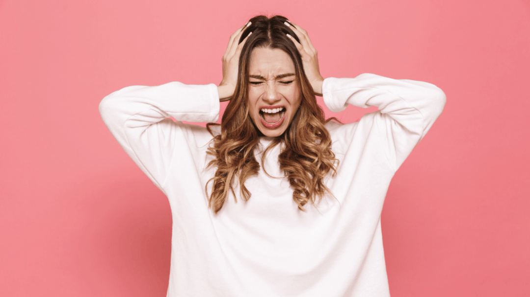 A frustrated young woman in a white sweater screaming with hands on her head against a pink background.