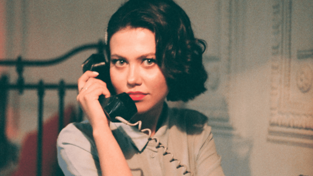 Woman with vintage hairstyle holding a black rotary phone, dressed in 1950s-style attire and looking directly at the camera.