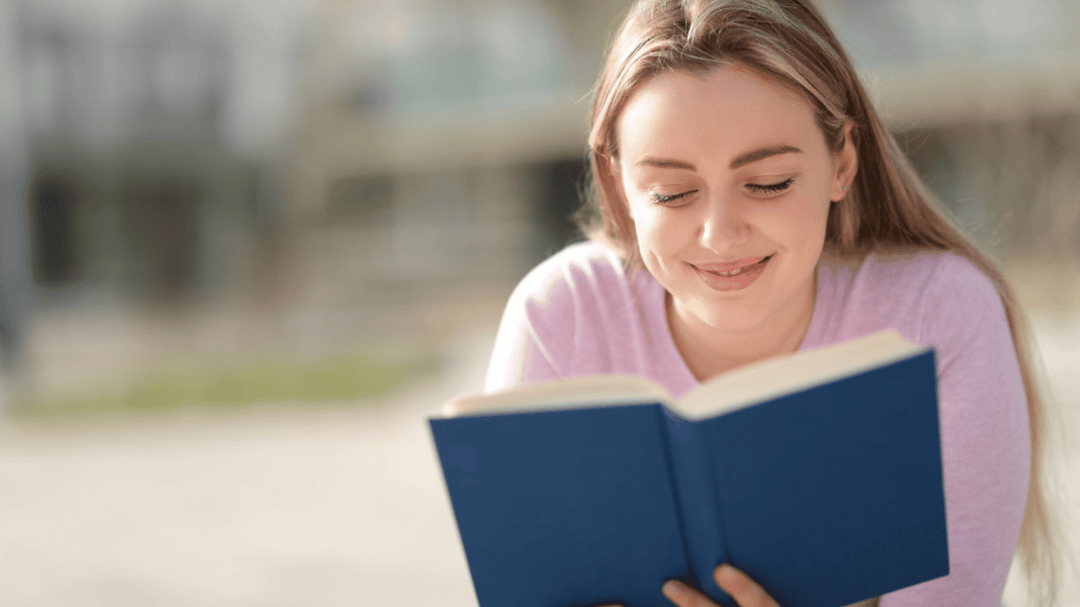 A young woman with long blonde hair smiles while reading a blue hardcover book outdoors, wearing a pink sweater.