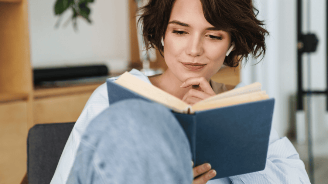 Woman with short brown hair wearing wireless earbuds, thoughtfully reading a book.