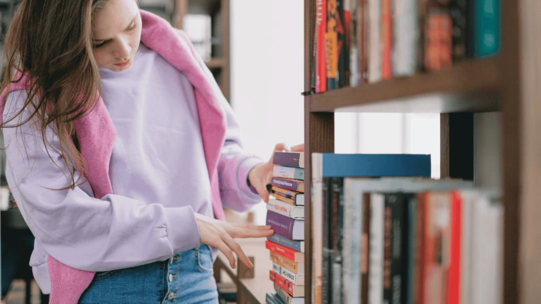 Photo of a woman browsing through books on a library shelf, wearing a light purple sweater and pink sweatshirt draped over shoulders.