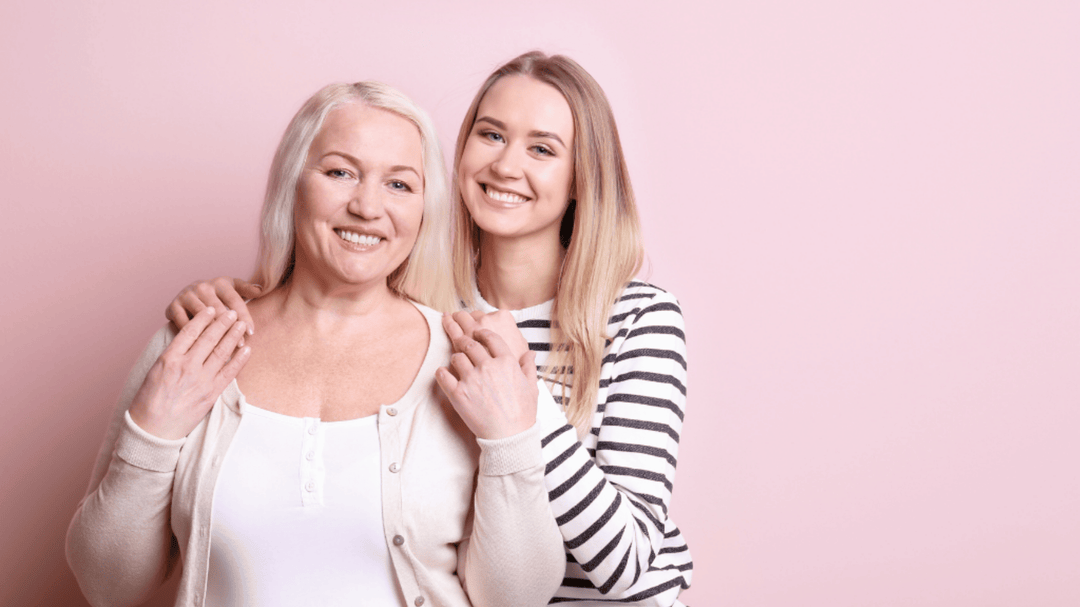 A smiling adult daughter stands behind her mother with both of them holding hands, posing in front of a soft pink background.