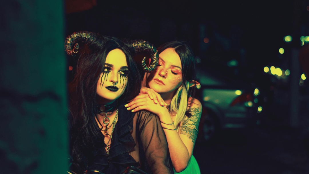 Two women in costume at night, one with horns and black makeup, the other with glitter and a dreamy expression on a motorcycle.