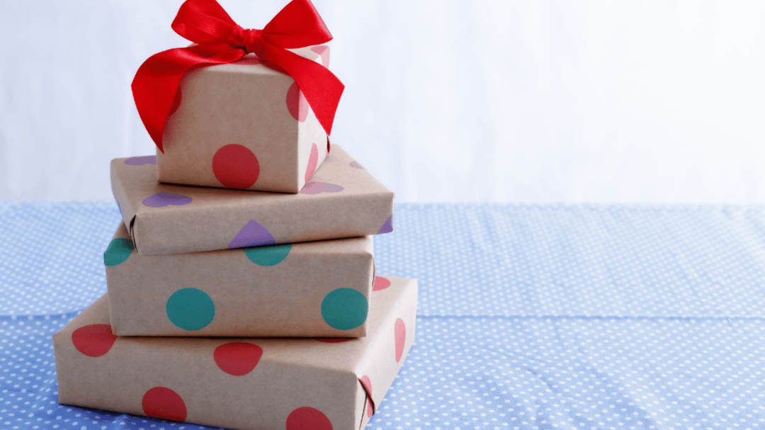 Stack of gift boxes wrapped in brown paper with colorful polka dots and topped with a red bow, placed on a blue polka dot tablecloth.