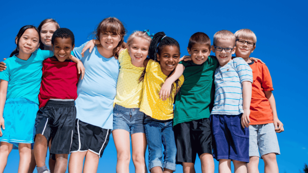 Group of smiling children standing in a row outdoors, arms around each other, against a bright blue sky.