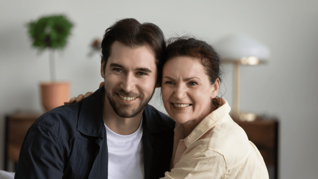 A smiling adult son and his mother sit close together on a couch, their arms wrapped around each other in a warm embrace.