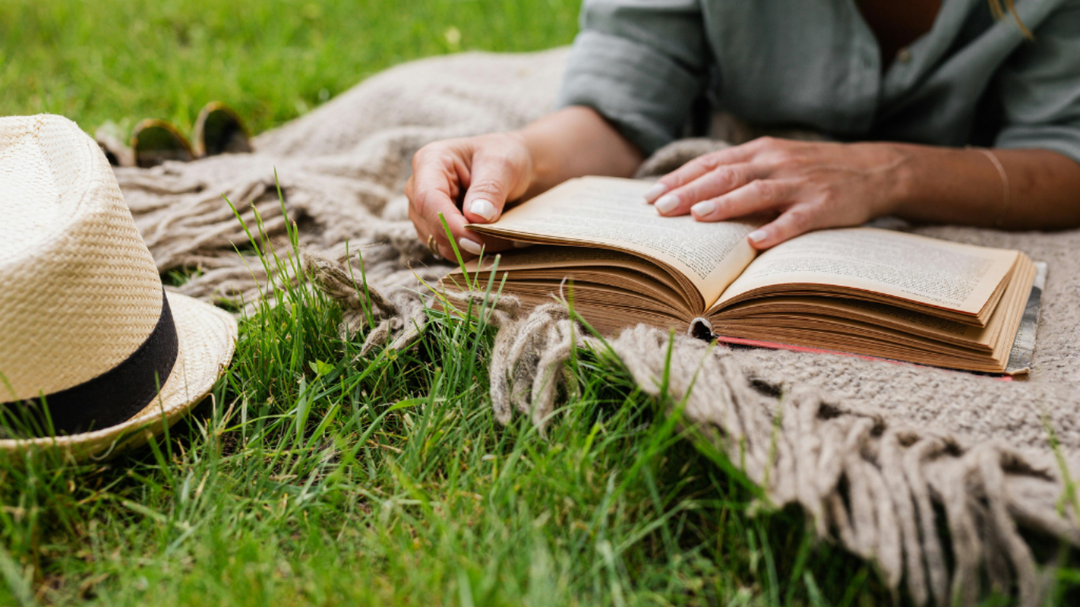 Person lying on a blanket in the grass reading an open book, with a straw hat placed nearby for a relaxed outdoor vibe.