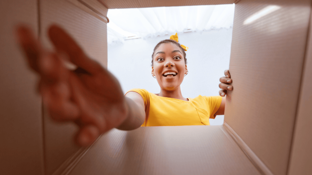A smiling girl reaches into a cardboard box, looking excited and surprised by what's inside.