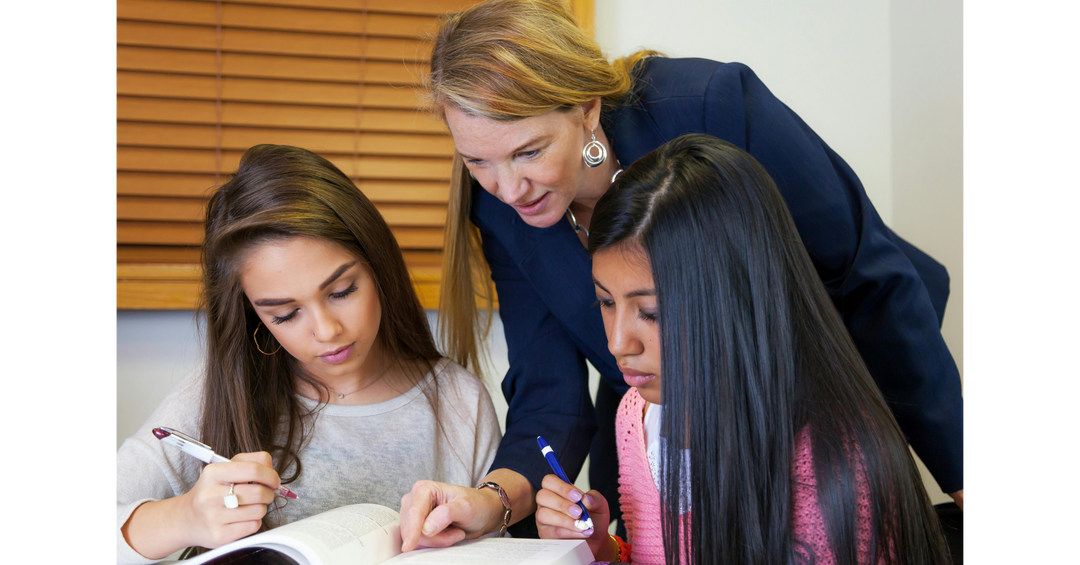 A teacher leaning over two students, guiding them as they read and write in a classroom setting.