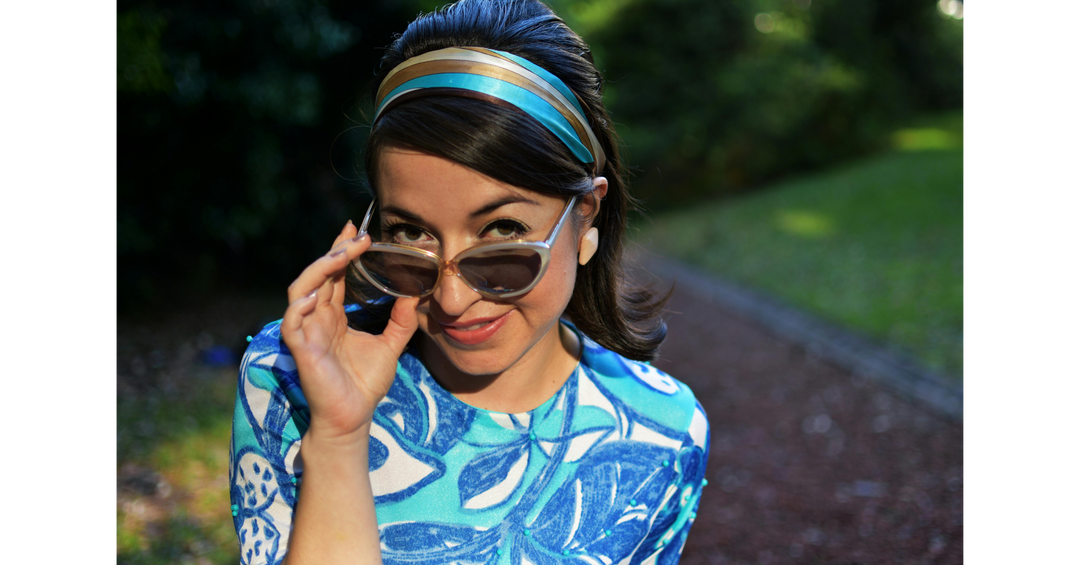 Woman wearing blue patterned dress and headband lifting sunglasses while standing on a leafy path outdoors.