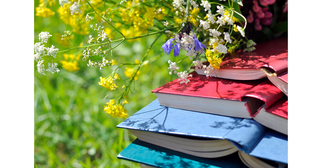 A colorful stack of hardcover books outdoors surrounded by wildflowers and bright summer greenery.