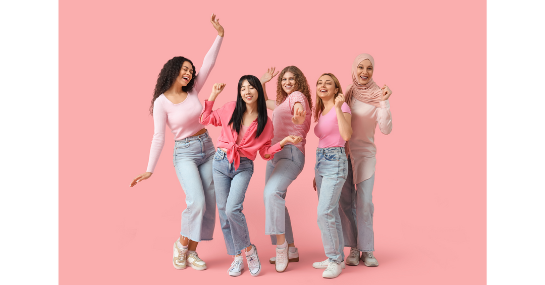 Diverse group of women dressed in pink outfits dancing together against a solid pink background.