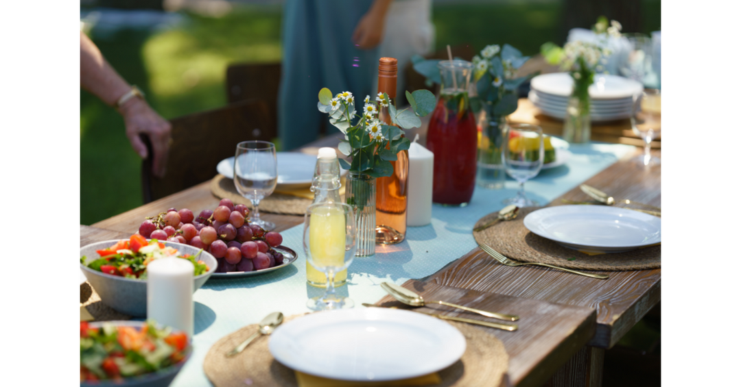 A rustic outdoor dining table is set with white plates, woven placemats, candles, flowers, fruit, salads, and colorful beverages.