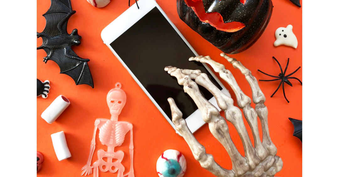 Smartphone surrounded by skeleton hands, bats, a spider, pumpkins, and Halloween decorations on a bright orange background.